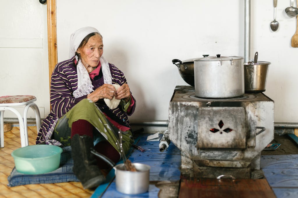 Aged ethnic female in headscarf sitting on floor near stove while kneading traditional bread at home