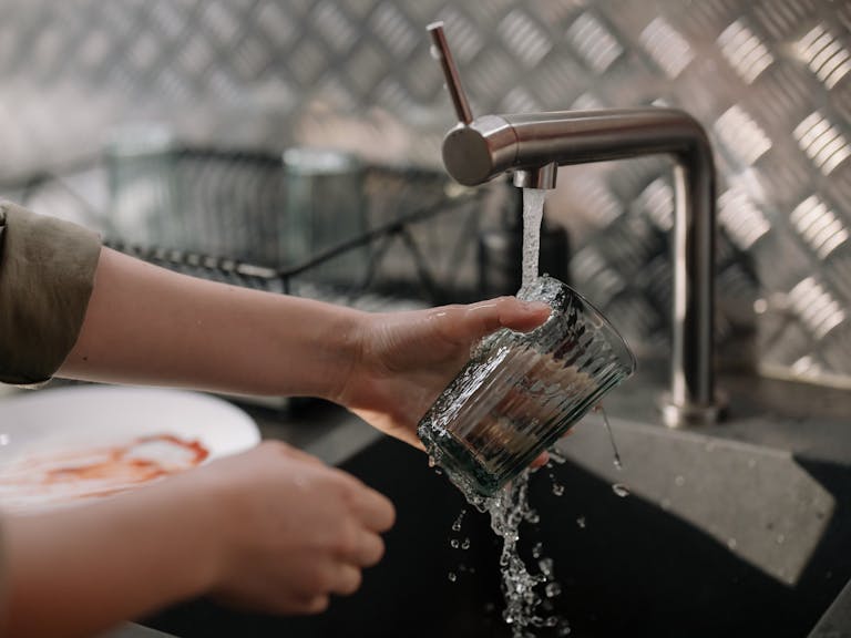 Hands washing a glass under running tap water in a home kitchen sink.